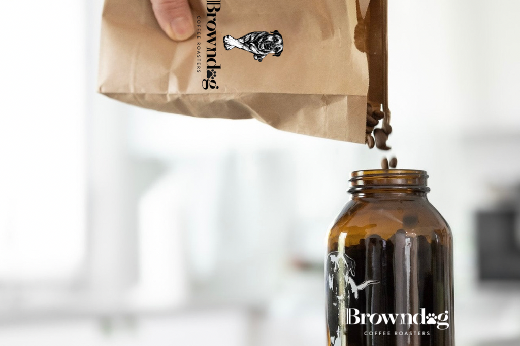 Hand pouring coffee from a Brownbag branded bag into an amber jar on a white surface.